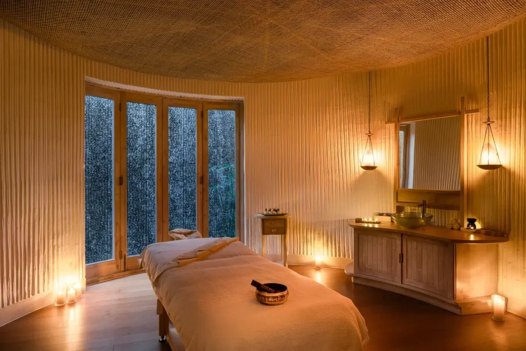 A softly lit treatment room contains a massage bed, timber walls, pendant lights, and tall windows facing greenery.