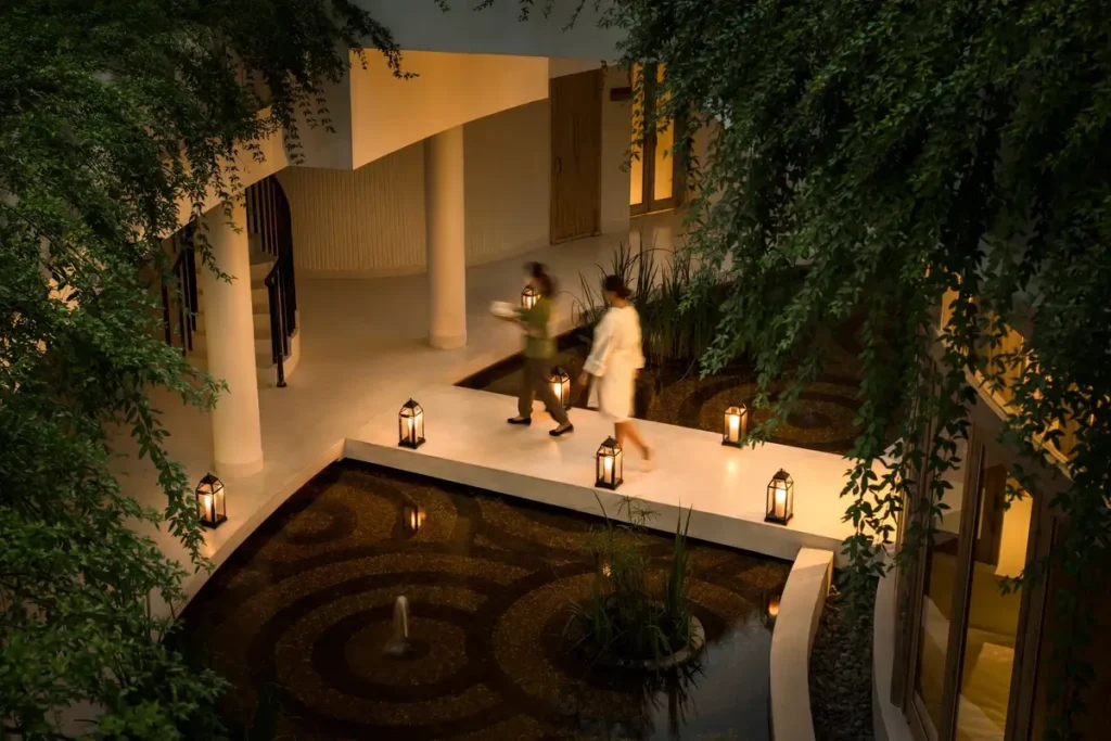 Two guests walk along a lantern-lined path beside a reflecting pool beneath trailing green plants.