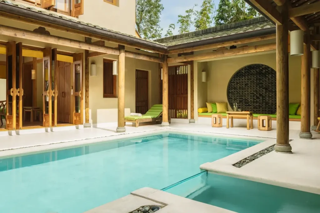 A turquoise private pool fills a courtyard enclosed by timber columns, pale walls, glass doors, and tiled roofs.