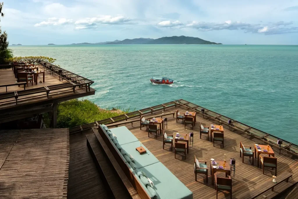 Dining tables occupy stepped wooden terraces above the sea, with an island on the horizon.