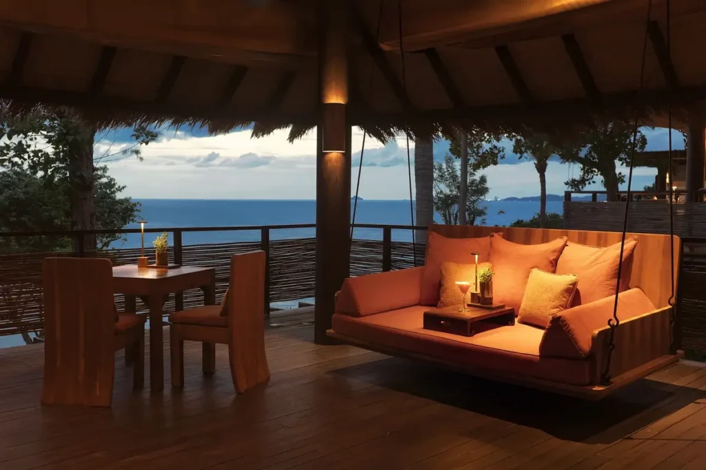 A cushioned wooden swing and dining table sit beneath a timber roof overlooking the sea at dusk.