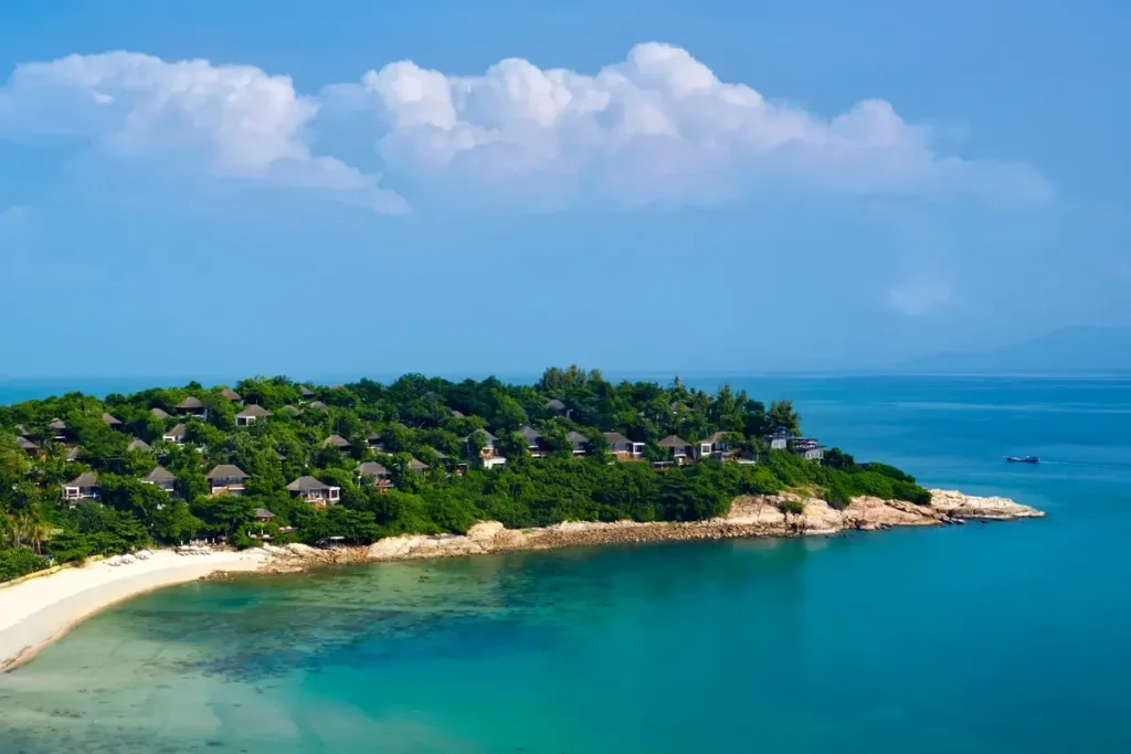 Villas nestle among trees on a rocky headland beside a curved sandy beach and turquoise sea at Six Senses Samui.