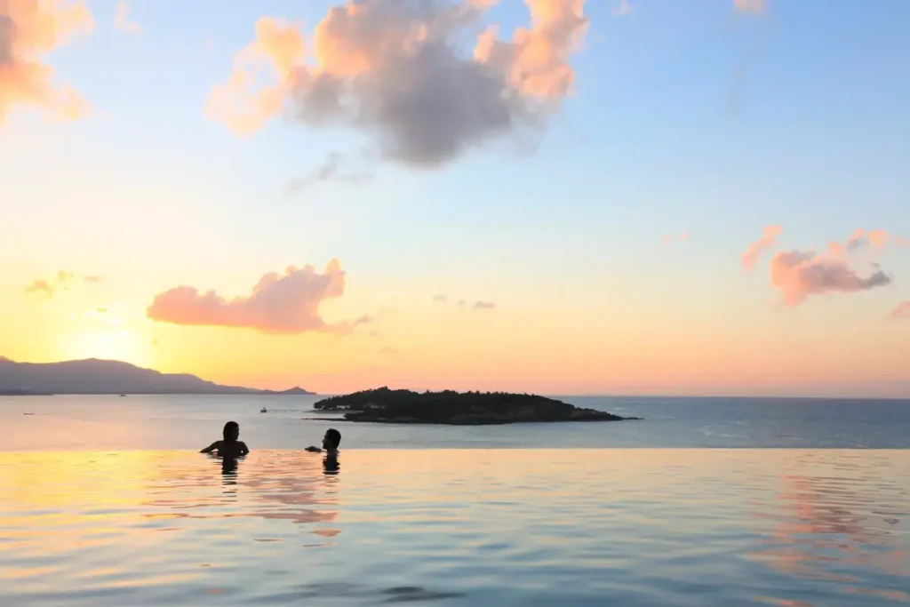 Two swimmers face the sea from an infinity pool beneath a pastel sunset sky, with an island beyond.