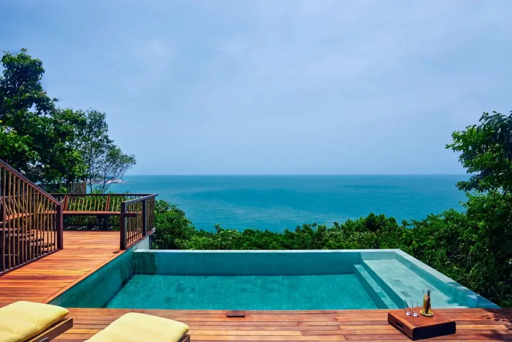 A rectangular infinity pool and wooden deck face the blue sea between tropical trees.