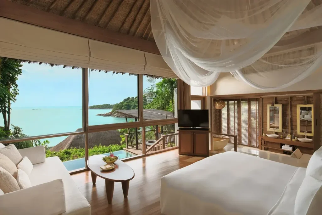A canopy-draped bed and pale sofa sit beneath a timber ceiling beside broad windows overlooking the sea.