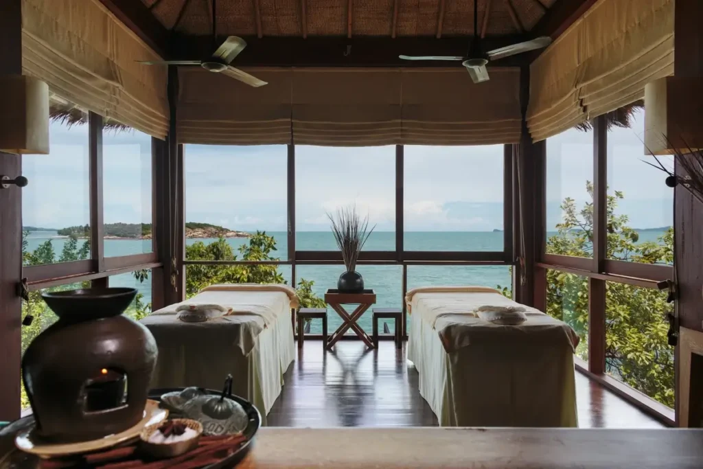 Two treatment tables stand in an open-sided timber spa room facing greenery and the sea.