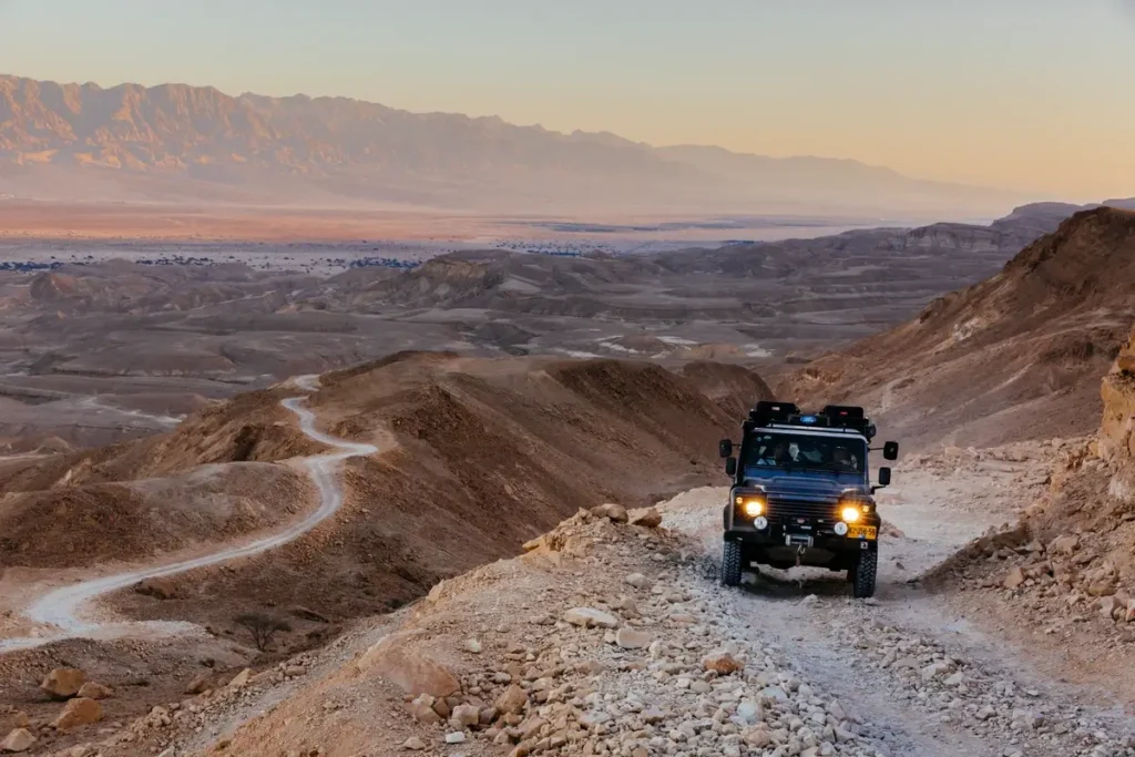 A dark off-road vehicle with headlights on follows a rocky track above a wide desert valley at dusk.