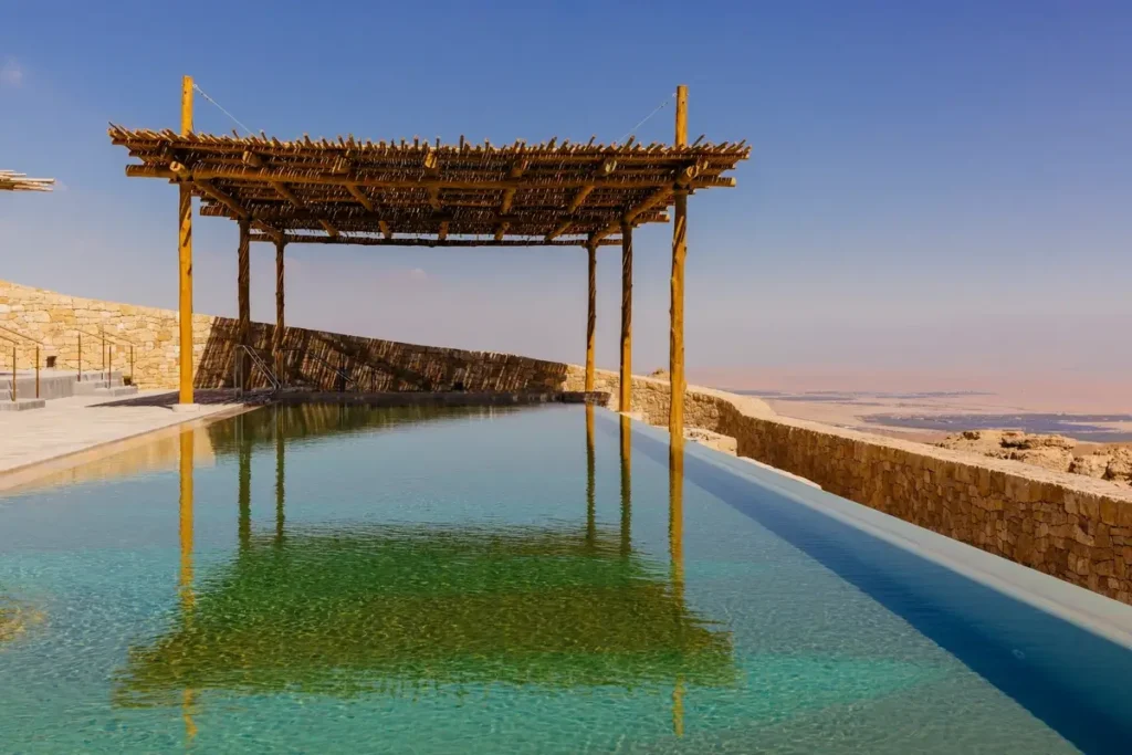 A long turquoise pool reflects a rustic pergola beside stone walls overlooking a pale desert valley.