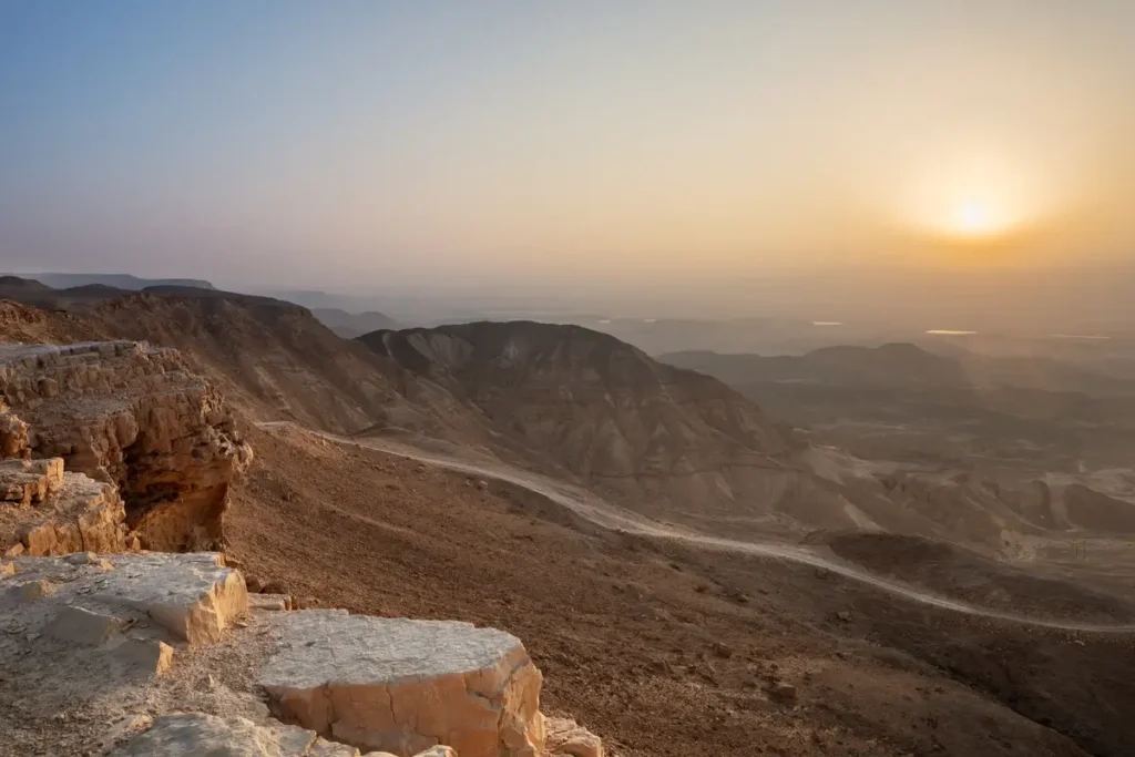The rising sun lights a rocky desert ridge, with layered mountains and a broad valley fading into haze.
