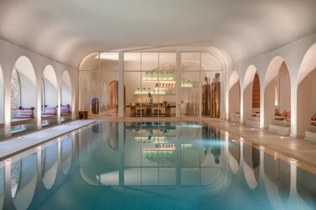A long indoor pool sits beneath a pale vaulted ceiling, with arched openings and loungers along its sides.