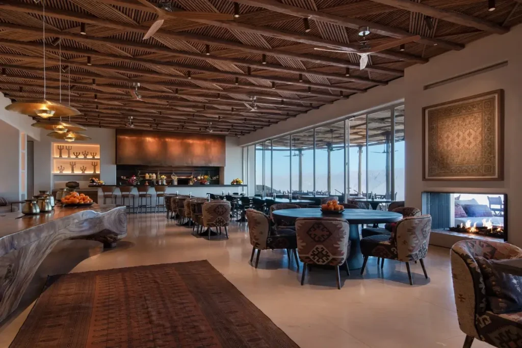 Tables and upholstered chairs sit beneath an exposed timber ceiling beside broad windows in Midian restaurant.