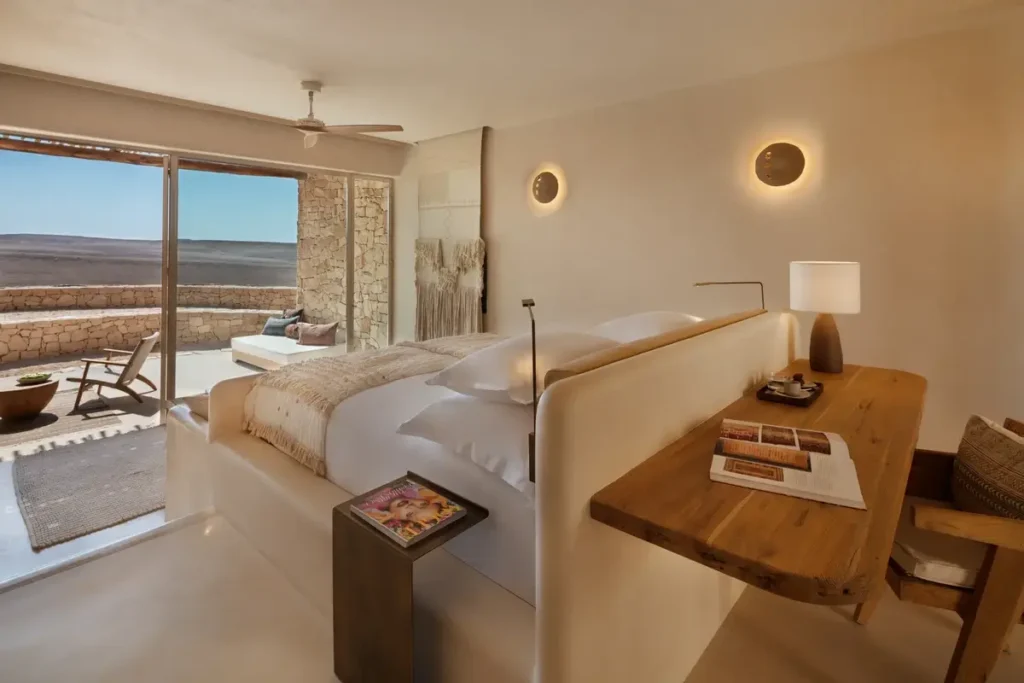 A pale bedroom with a wooden desk and circular wall lights opens through glass doors toward a desert-facing terrace.