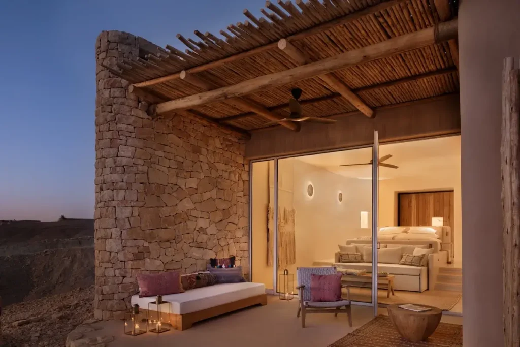 A stone-walled terrace with cushioned chairs and a daybed sits beneath a timber pergola beside a warmly lit bedroom.