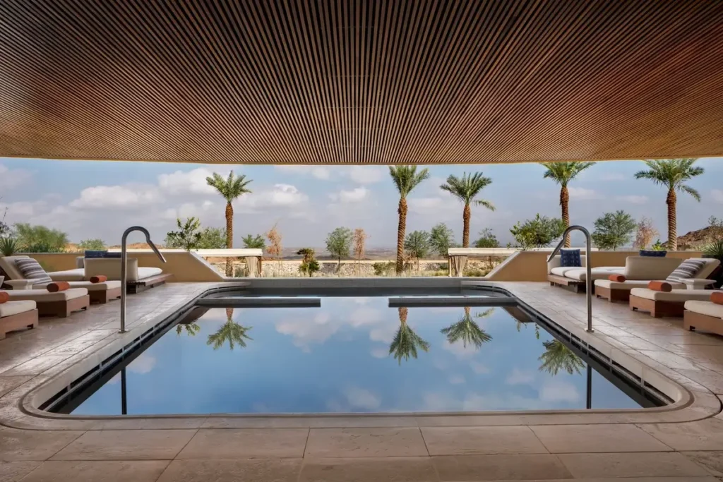 Rectangular spa pool beneath a timber ceiling with loungers, palms, and mountains beyond