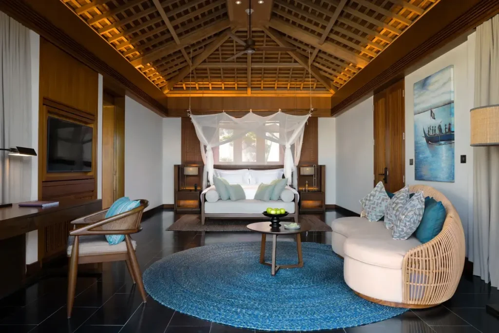 A timber-roofed villa interior contains a canopy bed, curved sofa, turquoise chairs, and a round blue rug.