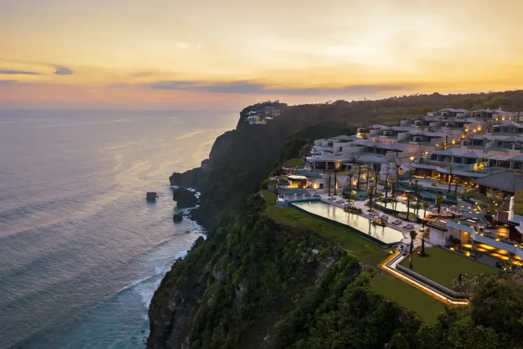 Resort buildings and illuminated terraces descend a steep green cliff above the ocean under a golden evening sky.