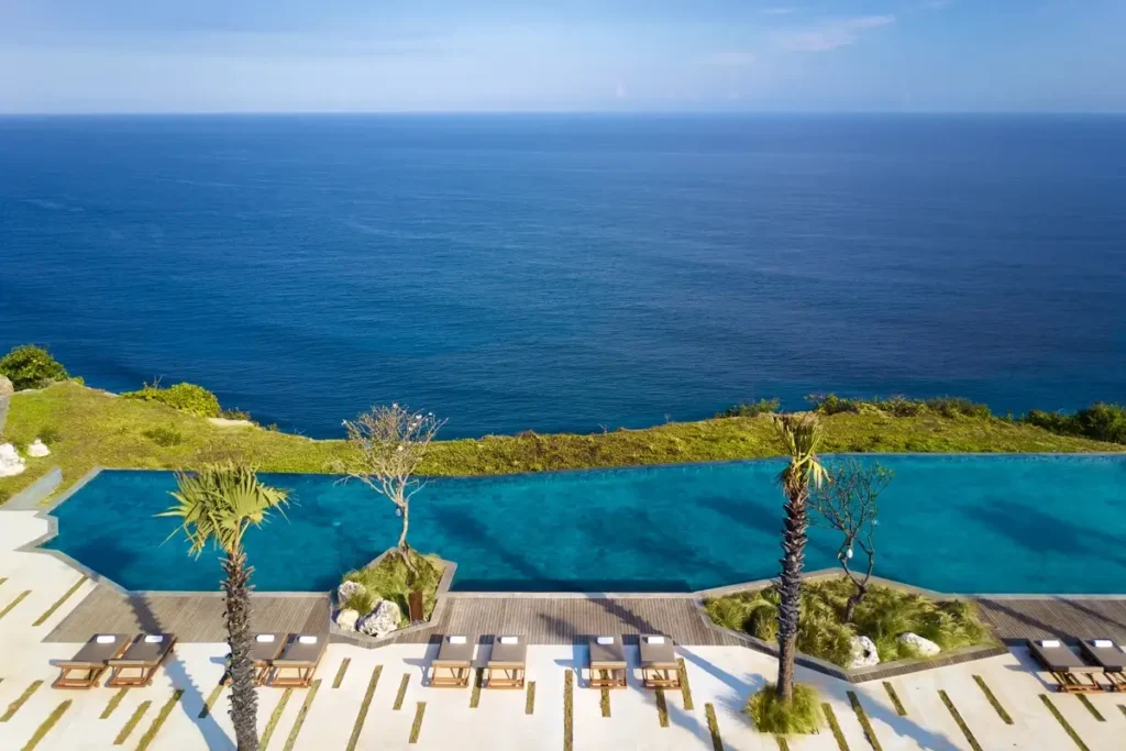 A curved infinity pool, palms, and rows of sun loungers overlook a wide expanse of blue ocean.