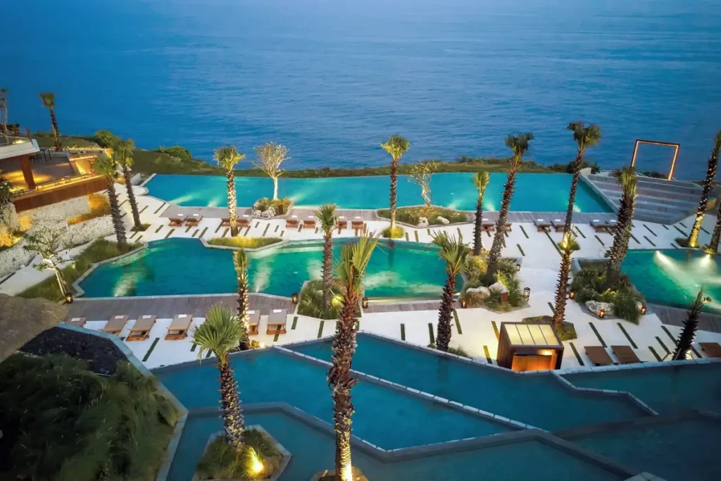Several turquoise pools step down landscaped terraces lined with palms and loungers toward the blue ocean.