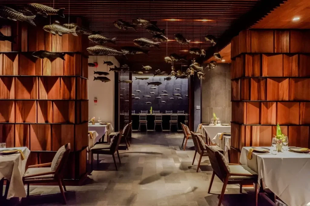 White-clothed tables sit between geometric timber columns beneath fish-shaped ceiling decorations in a dimly lit restaurant.