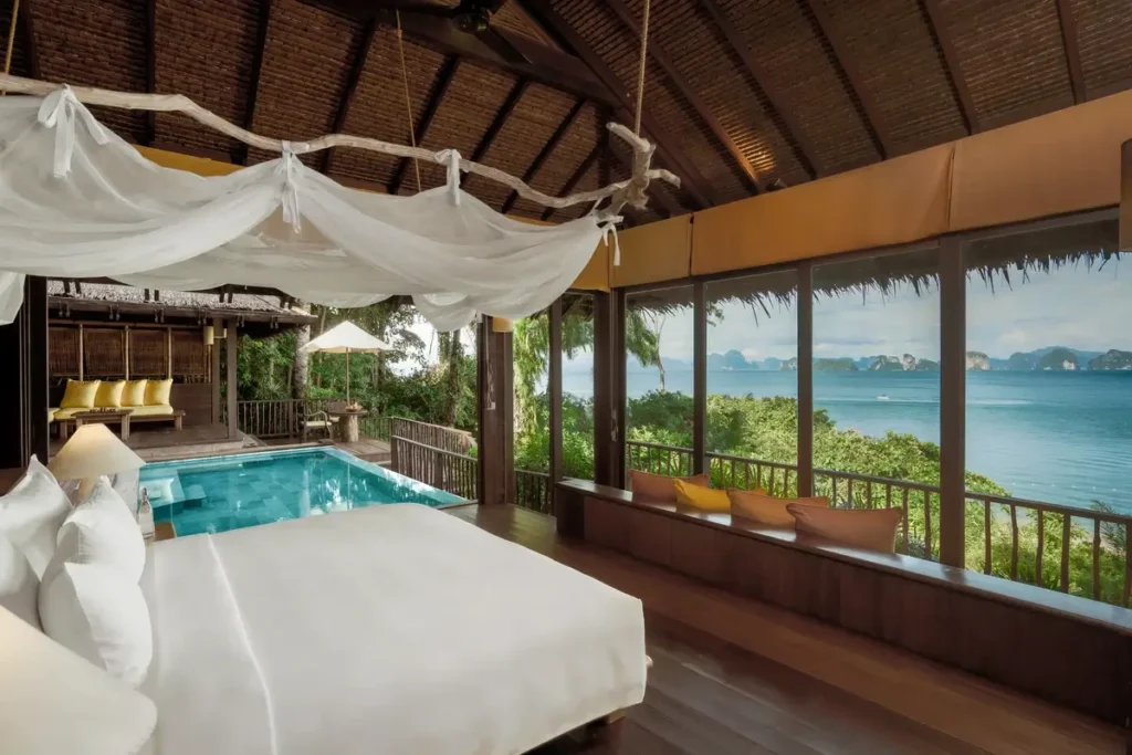 A timber bedroom with a canopy bed opens toward a private pool, with the sea visible through broad windows.