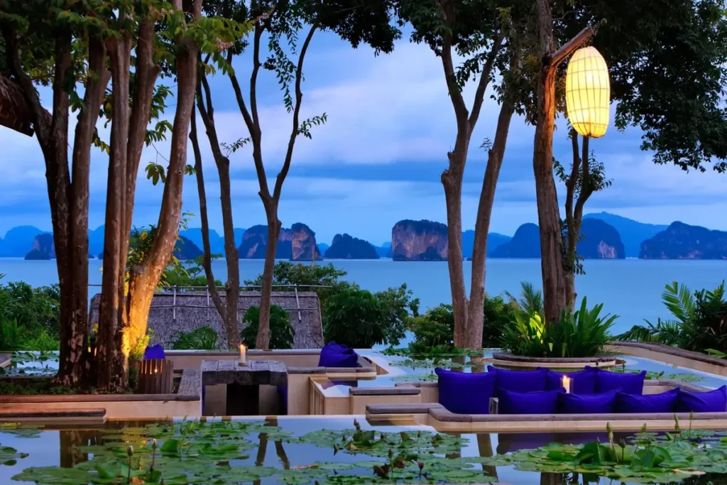 Purple cushions, timber platforms, and hanging lanterns furnish an outdoor lounge overlooking the bay at dusk.