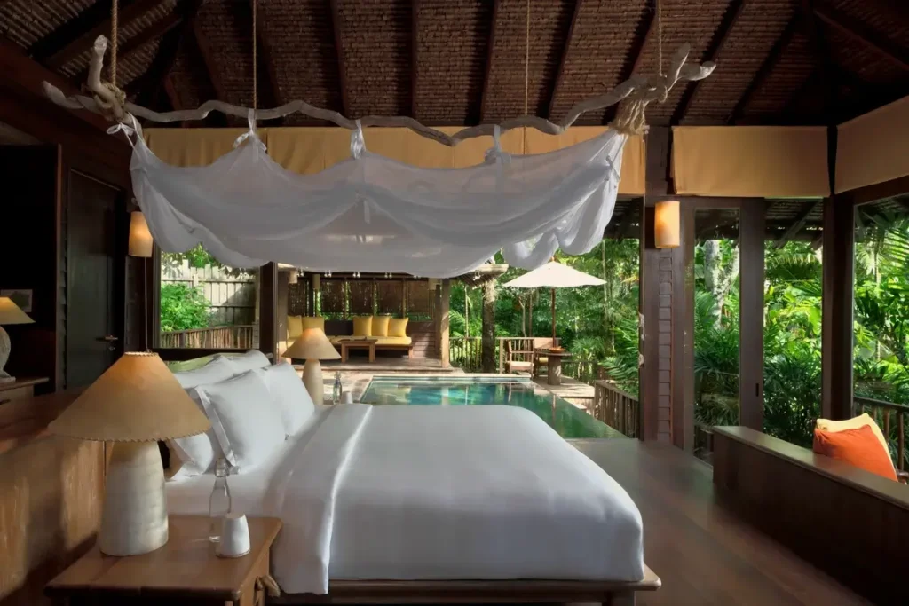 A canopy bed and timber rafters frame a rustic bedroom opening toward a private pool and tropical vegetation.