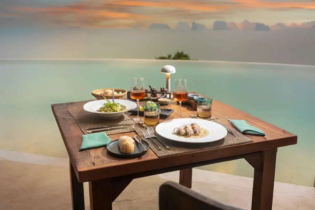 A wooden table set with food and drinks stands beside an infinity pool overlooking the bay at dusk.