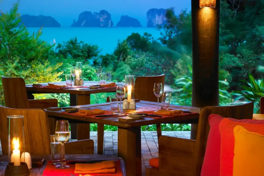 Wooden tables and chairs with colorful cushions sit among tropical plants overlooking the bay at blue hour.