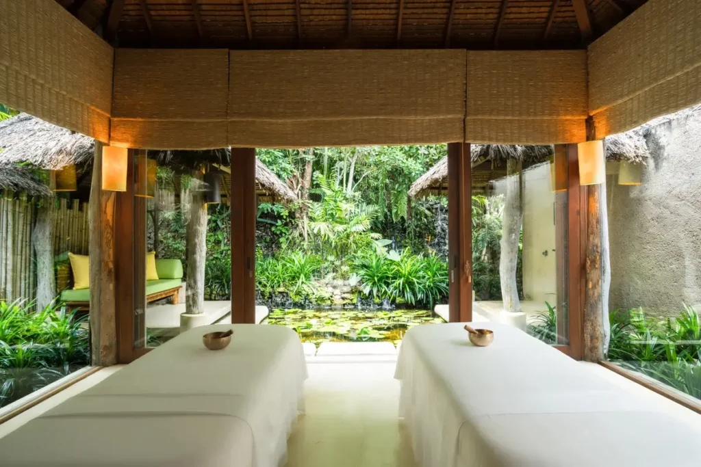 Two white treatment beds sit beneath a timber roof beside open blinds and a lush garden.