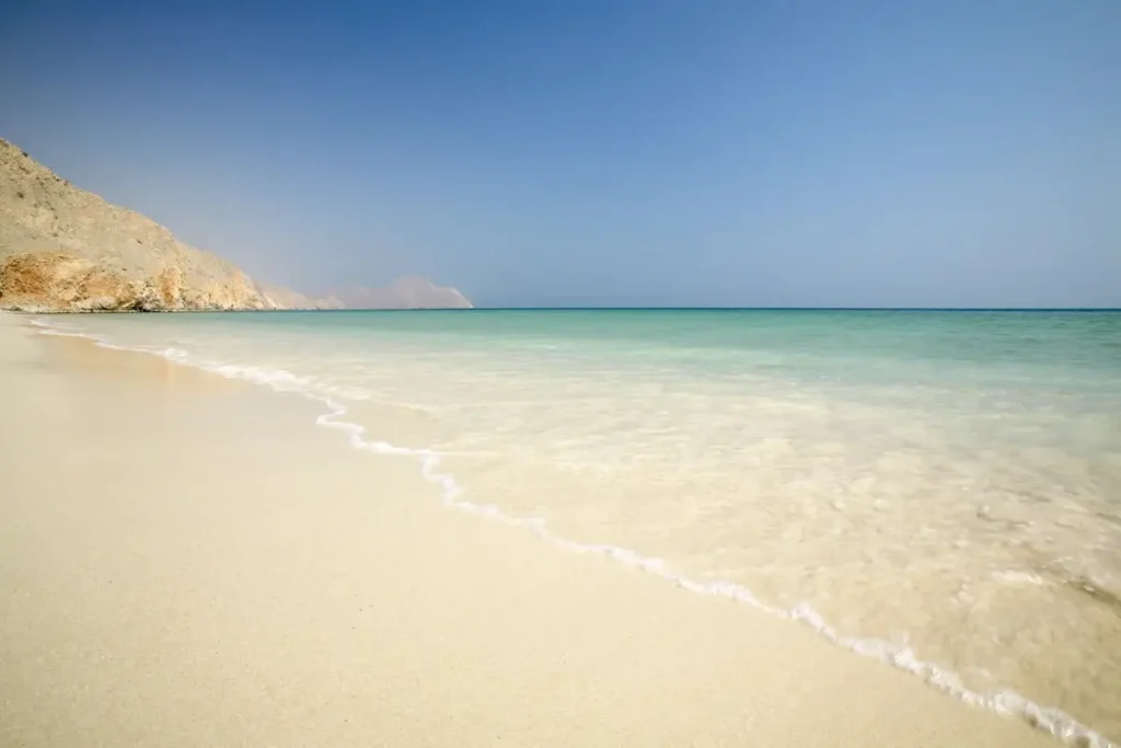 Pale sandy beach with gentle waves and a rocky mountain headland at Six Senses Zighy Bay