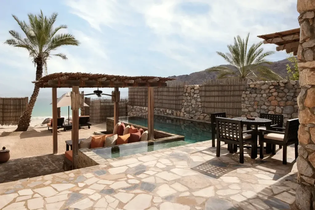 Stone terrace with a private pool, shaded daybed, dining table, and palms at Six Senses Zighy Bay