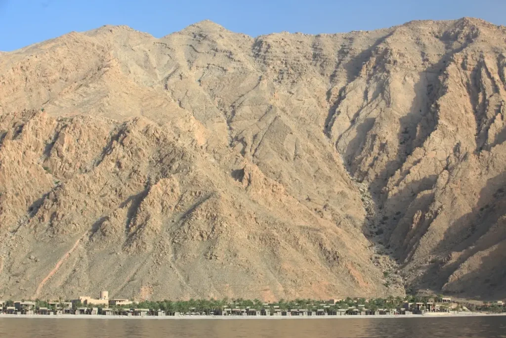 Rugged mountain slopes rising above a narrow strip of palms and resort buildings at Zighy Bay