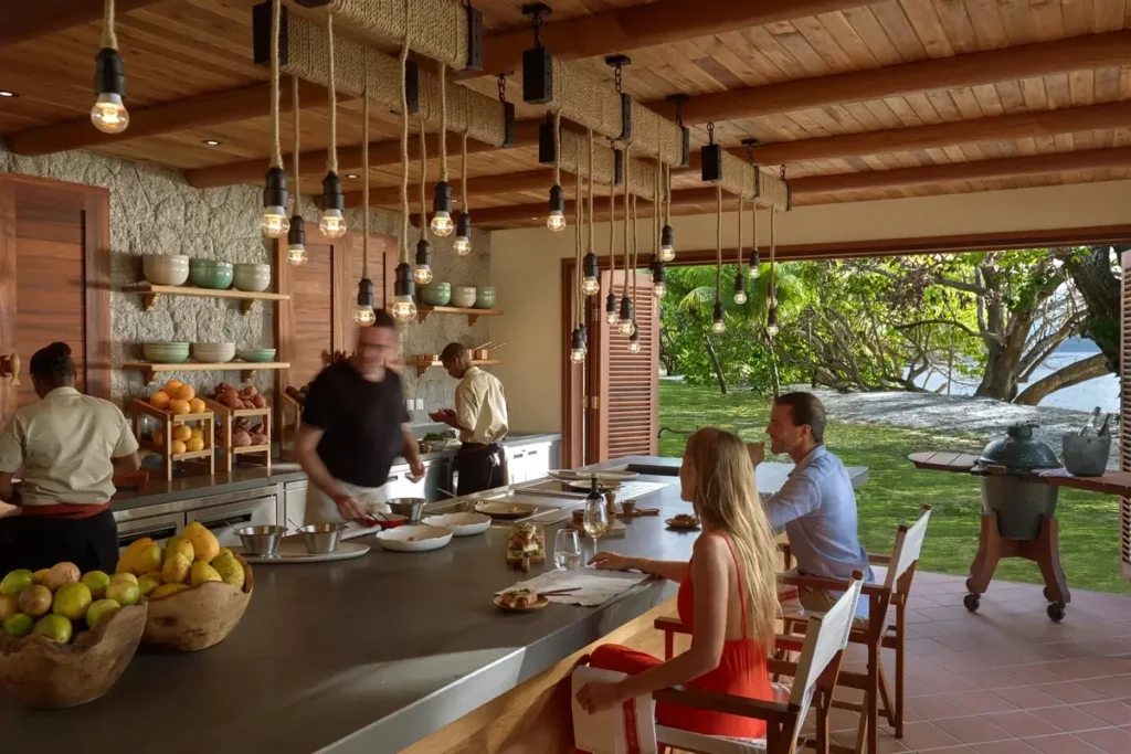 Guests sit at a kitchen counter while chefs prepare food beneath hanging lights in an open-sided timber pavilion.