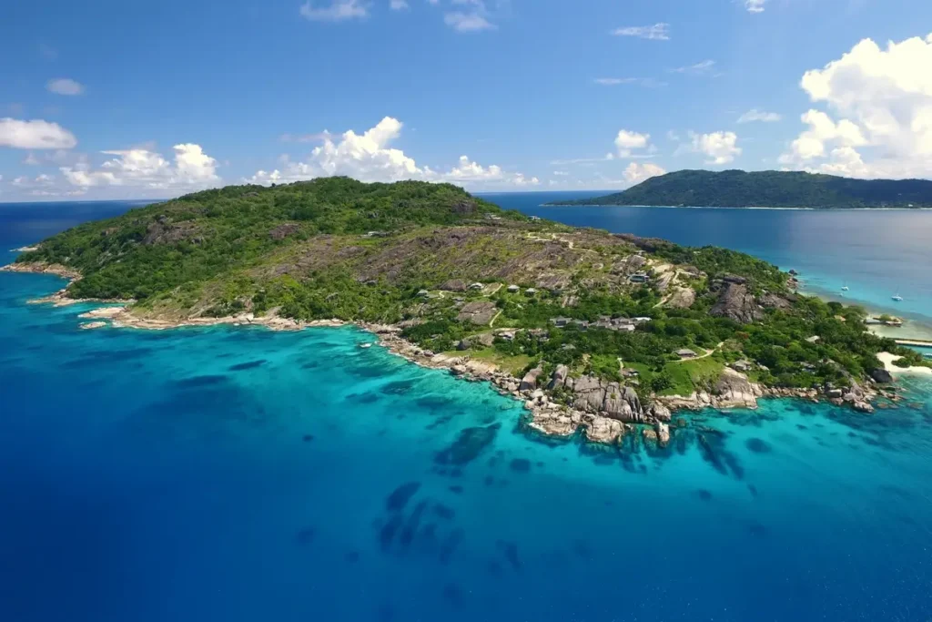 A green island with granite headlands is surrounded by turquoise shallows and deep blue ocean.