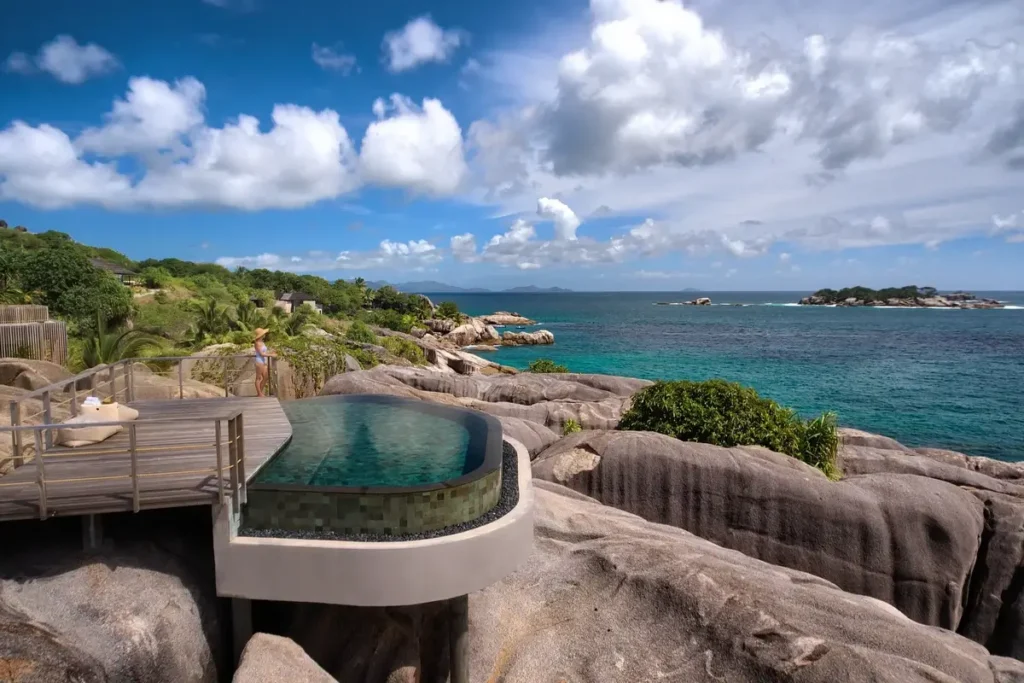A curved infinity pool and timber deck sit among large granite boulders overlooking the sea.