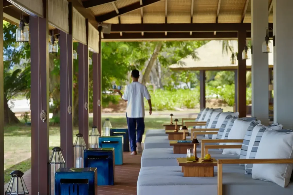 Pale cushioned chairs line a sheltered timber veranda with small tables, blue stools, and tropical greenery beyond.