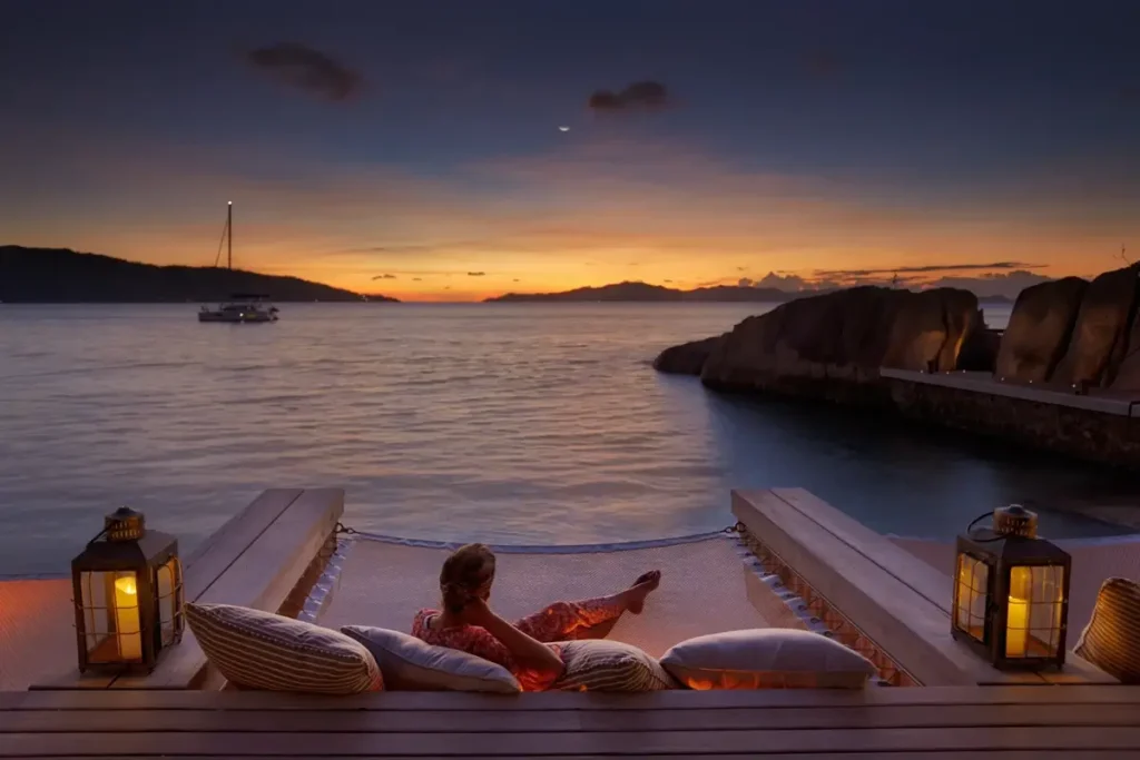 Guests recline on cushioned net seating above the water beside lanterns as an orange sunset lights the horizon.