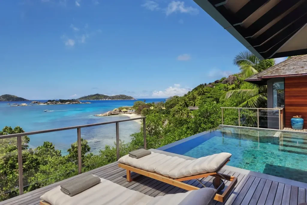 A padded lounger rests beside a private pool on a timber deck facing a blue bay and green coastal slopes.