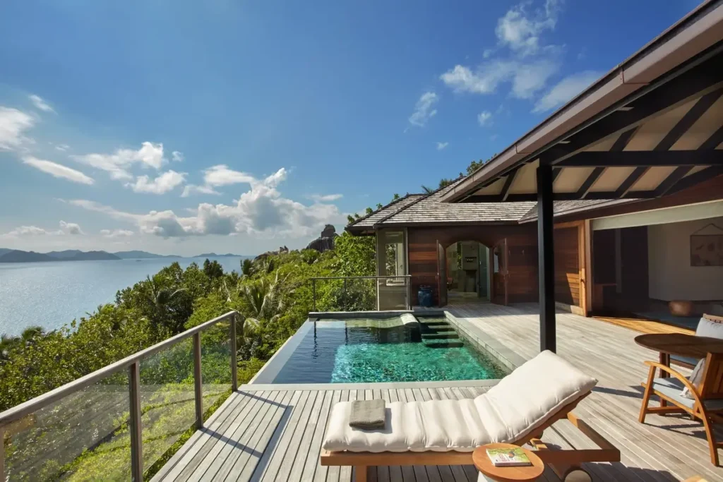 A timber deck with a cushioned lounger and private infinity pool overlooks trees and the ocean from a hillside villa.